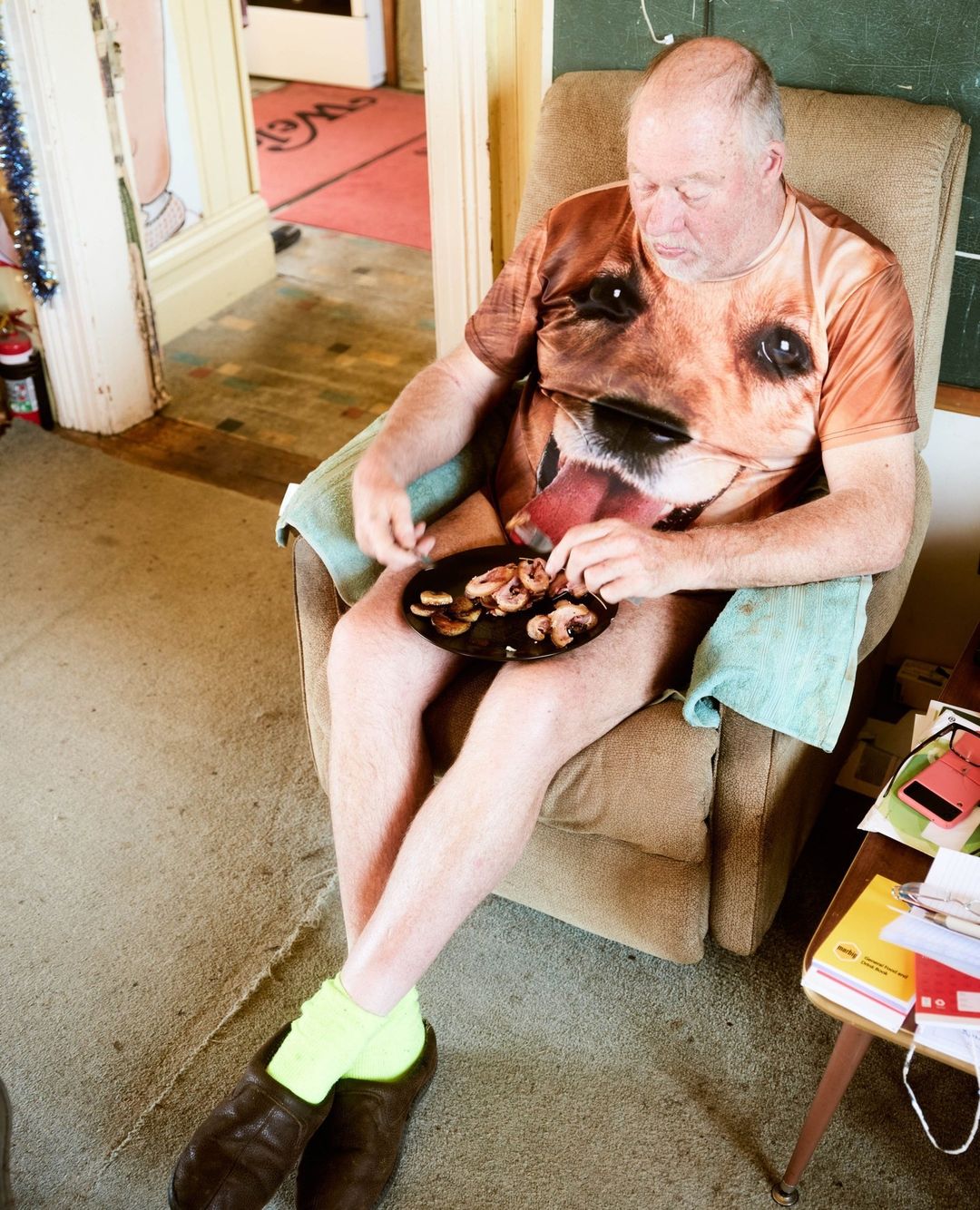 An older man in a dog-print shirt eats from a plate while sitting in a tan armchair with oversized shoes indoors.