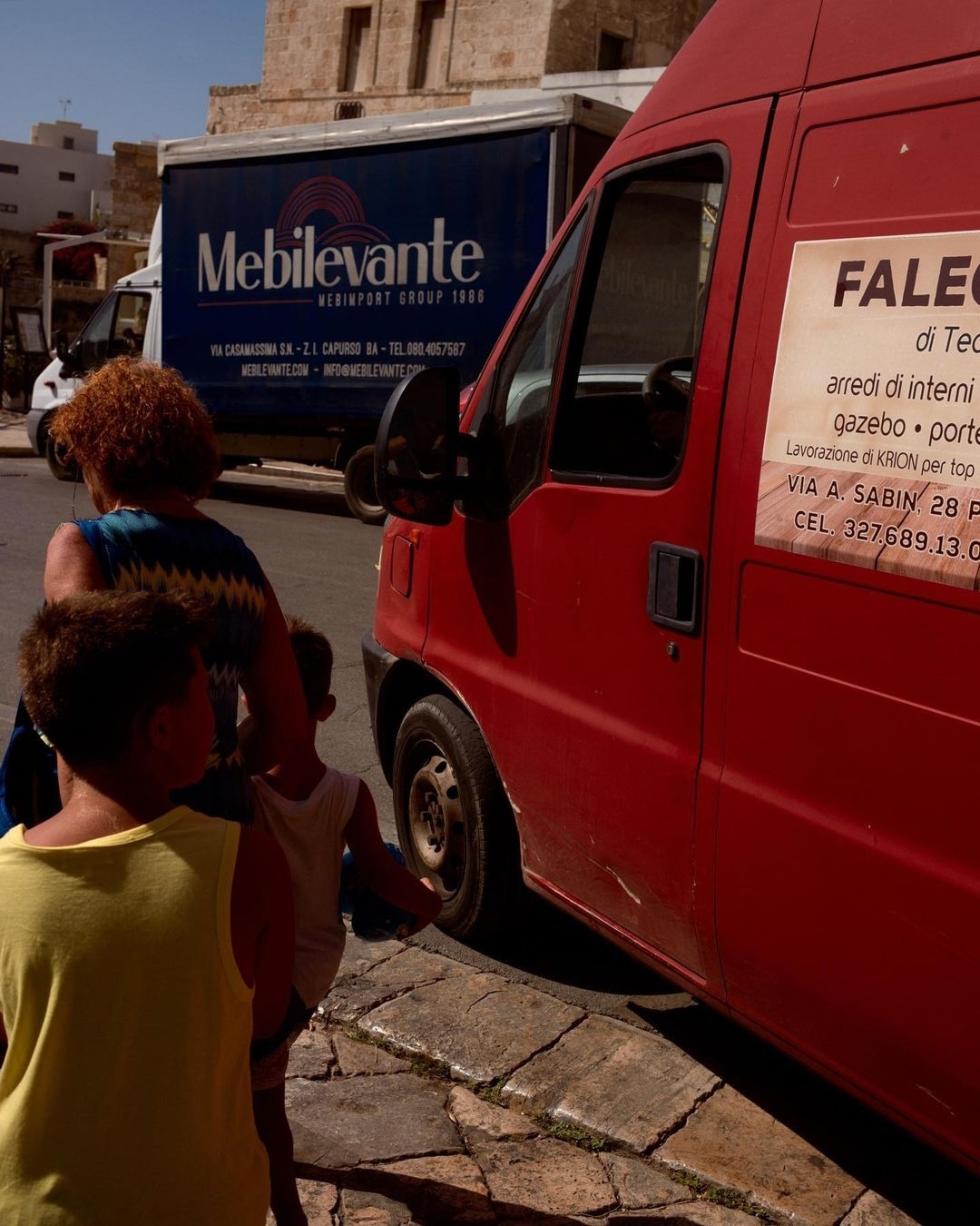 Family walking by red delivery van with blue shipping truck and old buildings in the background on a sunny day in an urban area.