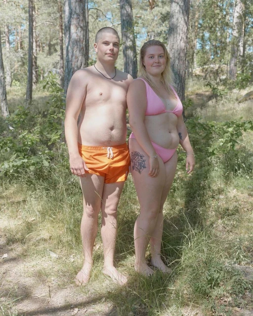 Couple poses in a forest. A shirtless man with orange swim trunks stands next to a woman in a pink bikini in a lush wooded area.