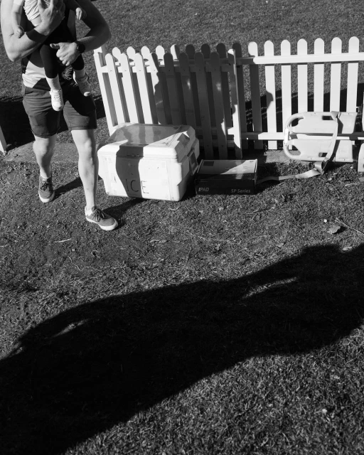 Monochrome shot of an adult carrying a child on grass next to a small white fence and various boxes on a sunny day.