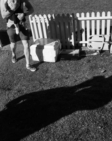 Monochrome shot of an adult carrying a child on grass next to a small white fence and various boxes on a sunny day.