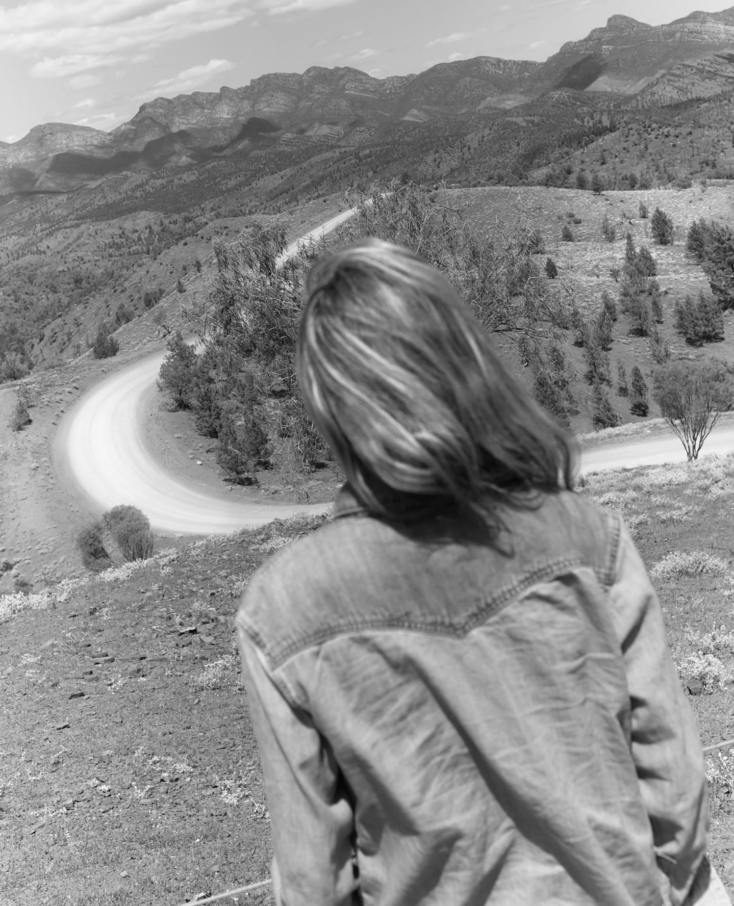 Monochrome landscape featuring a woman in a denim shirt overlooking a winding road and distant mountains under a cloudy sky.