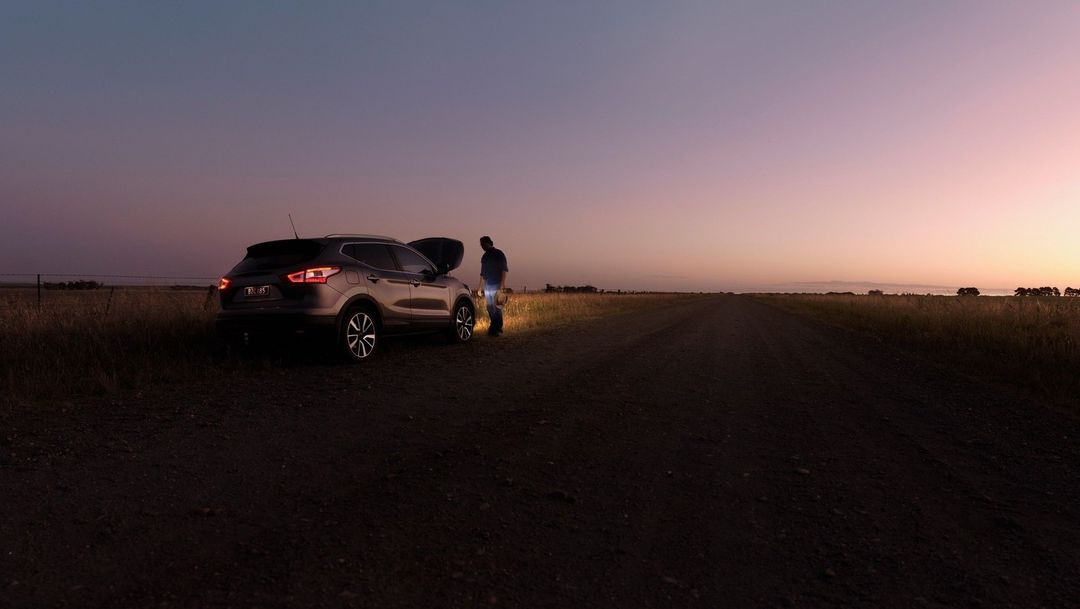 Man working on car engine with open hood on rural road at sunset. Car tail lights are on. Field and sky in background.