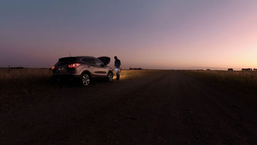 Man working on car engine with open hood on rural road at sunset. Car tail lights are on. Field and sky in background.