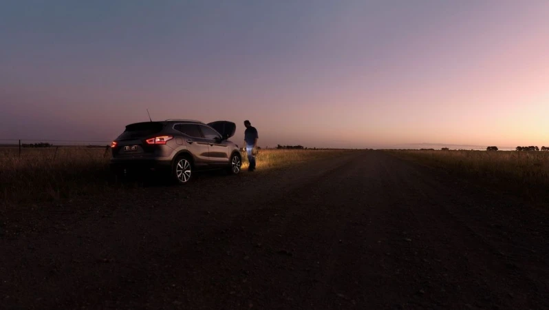 Man working on car engine with open hood on rural road at sunset. Car tail lights are on. Field and sky in background.
