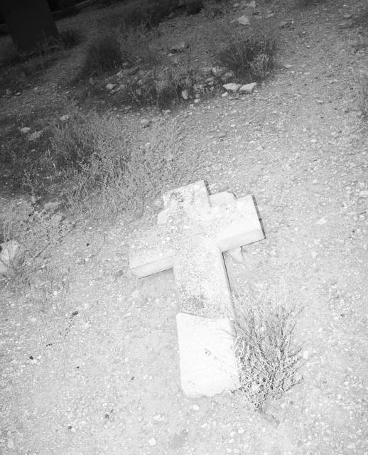 A weathered stone cross lies on a gravelly surface with sparse vegetation, captured in a black and white, high-angle shot.