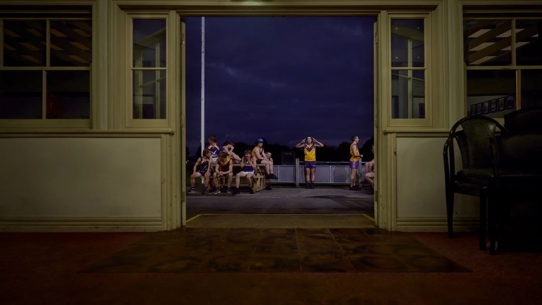 A group of young football players sits and stands outside a building with windows and open doors, set against a night sky.