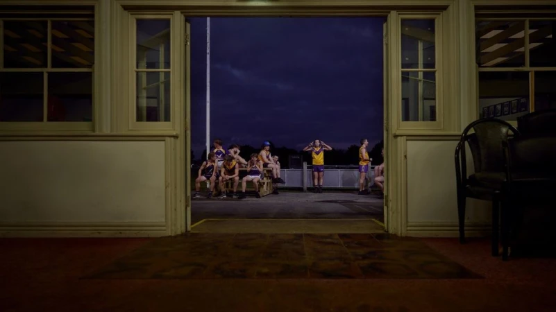 A group of young football players sits and stands outside a building with windows and open doors, set against a night sky.