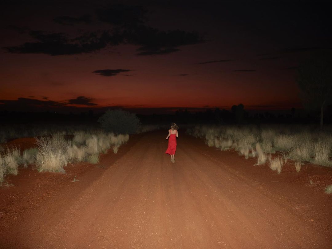 Woman in red dress runs down dirt road at dusk, with dry grass and dark red sky above, creating a scene of solitude and movement.