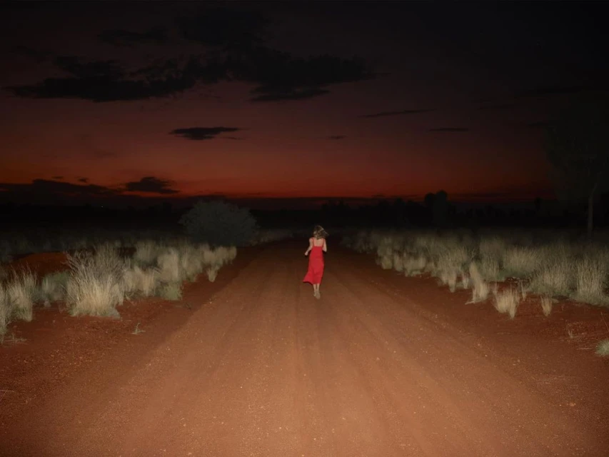 Woman in red dress runs down dirt road at dusk, with dry grass and dark red sky above, creating a scene of solitude and movement.