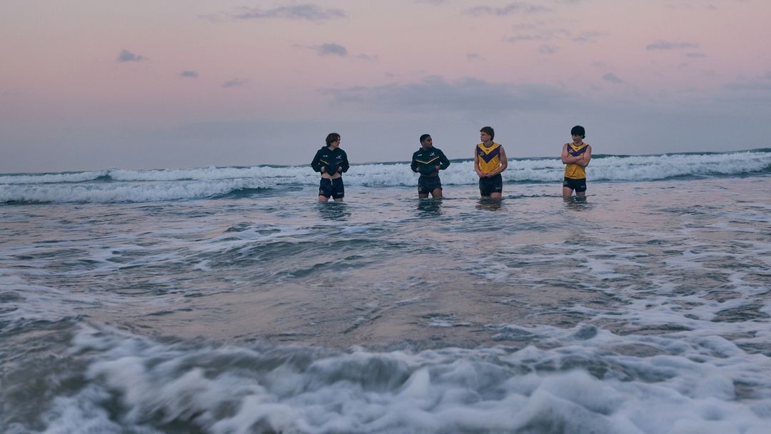 Four people stand waist-deep in ocean waves at dusk. They wear athletic wear and face forward as the waves come toward the viewer.