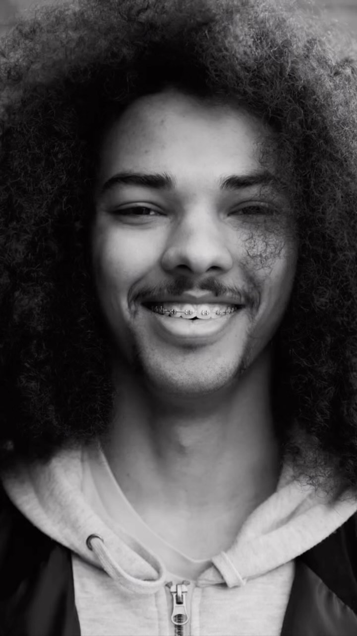 Close-up portrait of a young adult male with curly afro, wearing a hoodie, looking at the camera with a slight smile, black and white.