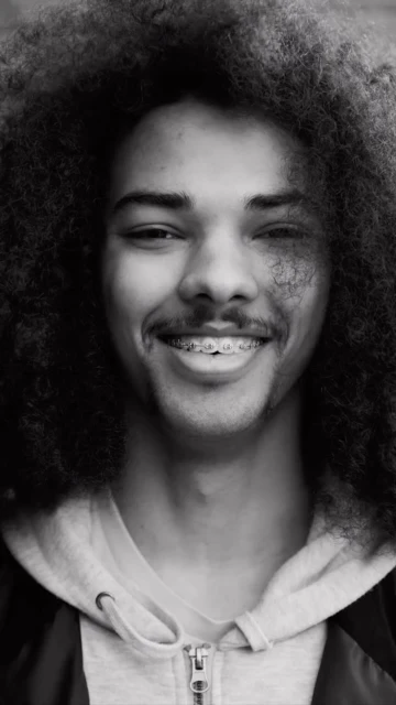 Close-up portrait of a young adult male with curly afro, wearing a hoodie, looking at the camera with a slight smile, black and white.