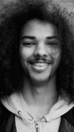 Close-up portrait of a young adult male with curly afro, wearing a hoodie, looking at the camera with a slight smile, black and white.