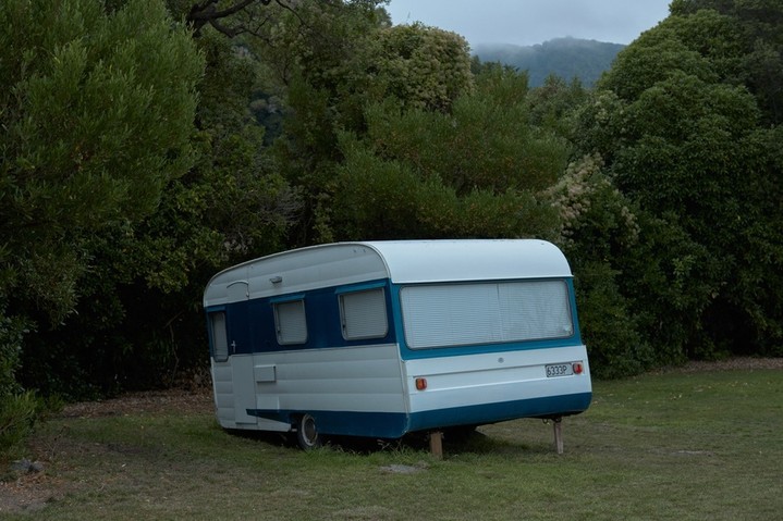 White and blue vintage caravan parked on green grass near a dense green forest. Overcast sky and muted colors.