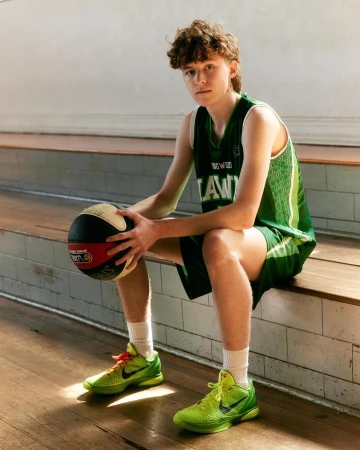 Young man in a green basketball uniform holding a basketball while seated on wooden bleachers