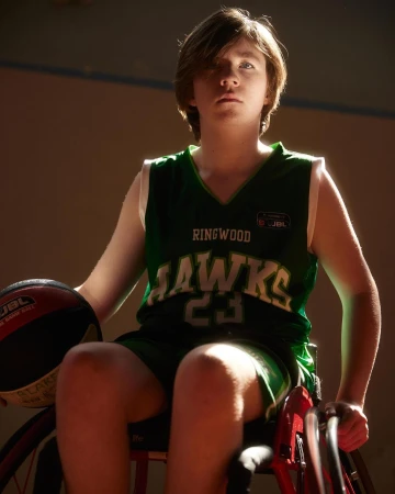 Young athlete in a green basketball jersey holding a basketball while seated in a sports wheelchair indoors
