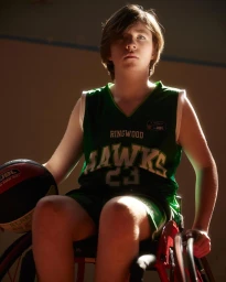 Young athlete in a green basketball jersey holding a basketball while seated in a sports wheelchair indoors