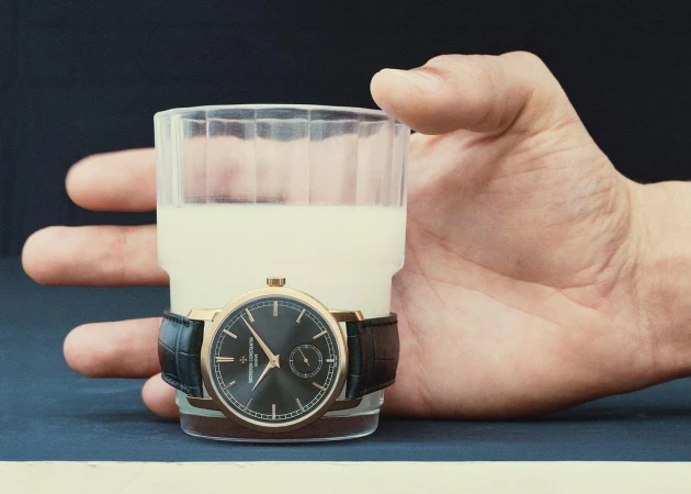 Close-up of a clear glass of white liquid, a luxury wristwatch on the glass, and a partial view of an adult human hand against a dark backdrop.
