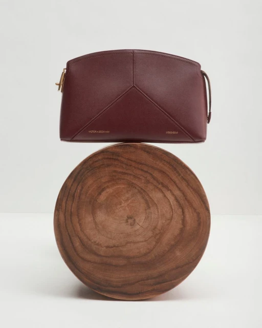 Burgundy leather handbag on a wooden stand against a plain white background in a studio-style product shot. Minimalist aesthetic.