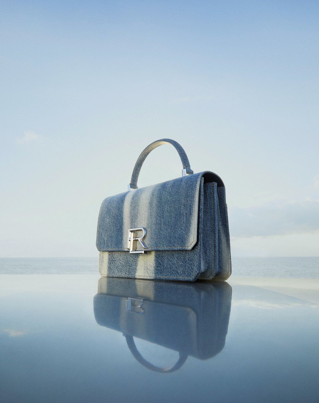 Blue denim handbag with silver clasp sits on reflective surface against pale blue sky and horizon in fashion product shot.
