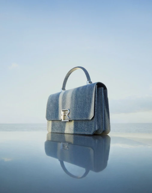 Blue denim handbag with silver clasp sits on reflective surface against pale blue sky and horizon in fashion product shot.