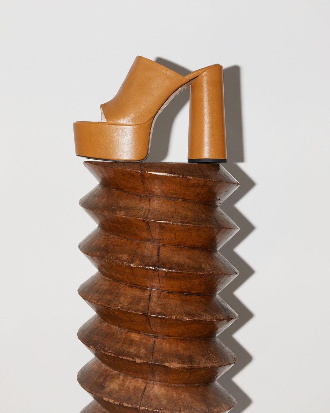 Studio shot of a single tan platform mule on top of a carved wooden stand against a white background in a vertical composition.