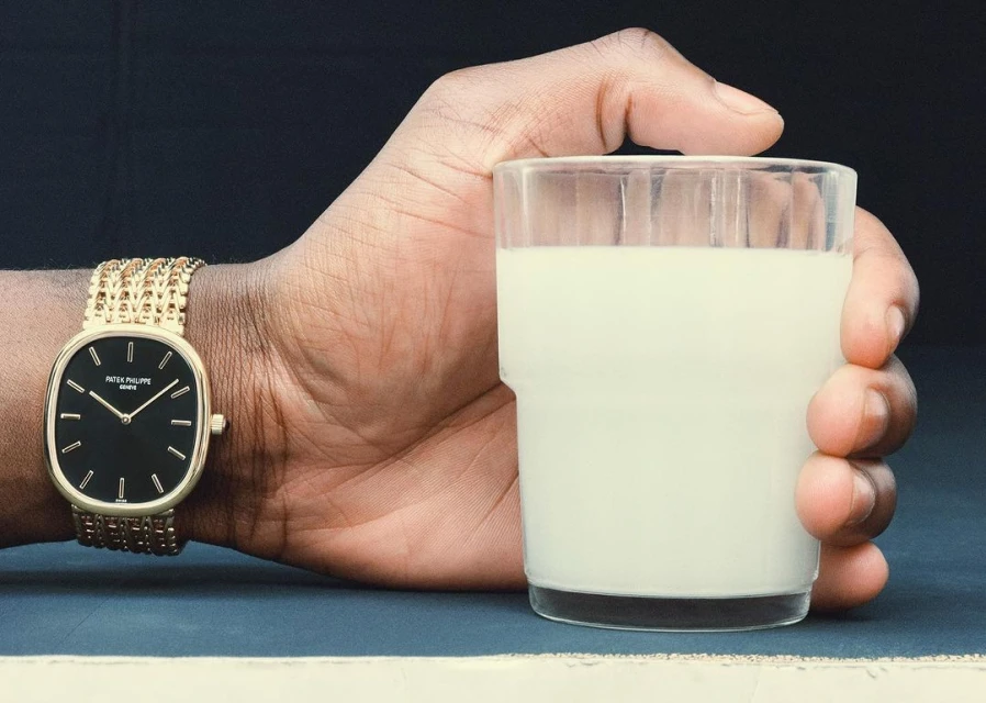 Close-up of a dark-skinned hand holding a glass of milk and wearing a gold Patek Philippe watch against a dark background.