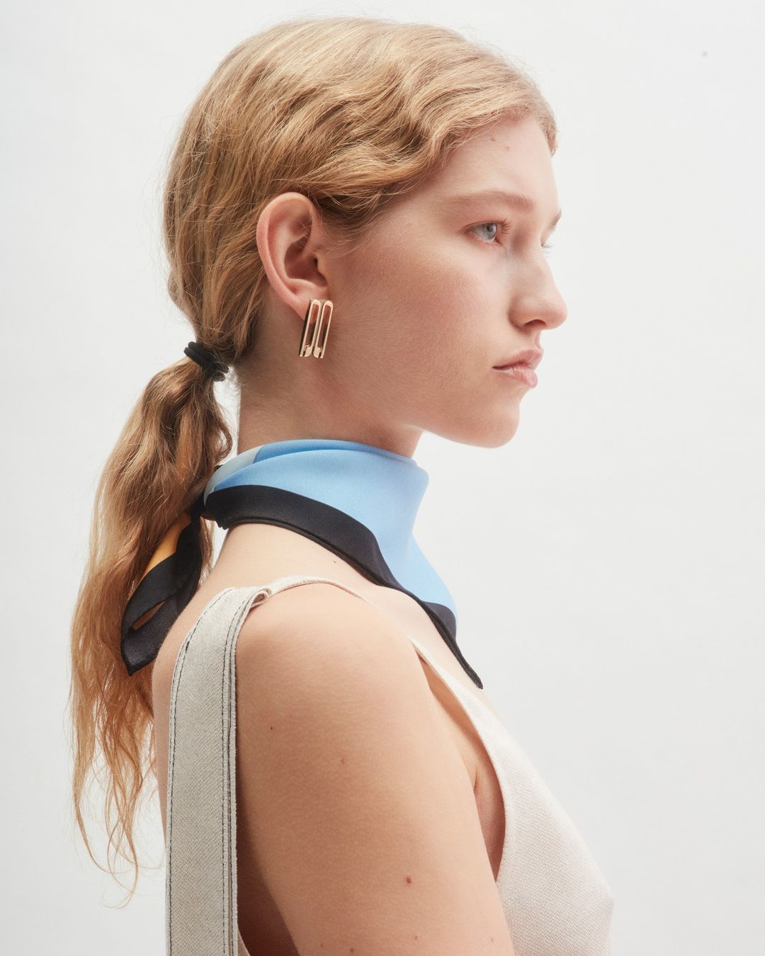 Profile portrait of a woman wearing a blue and black silk scarf and gold earrings against a plain white background