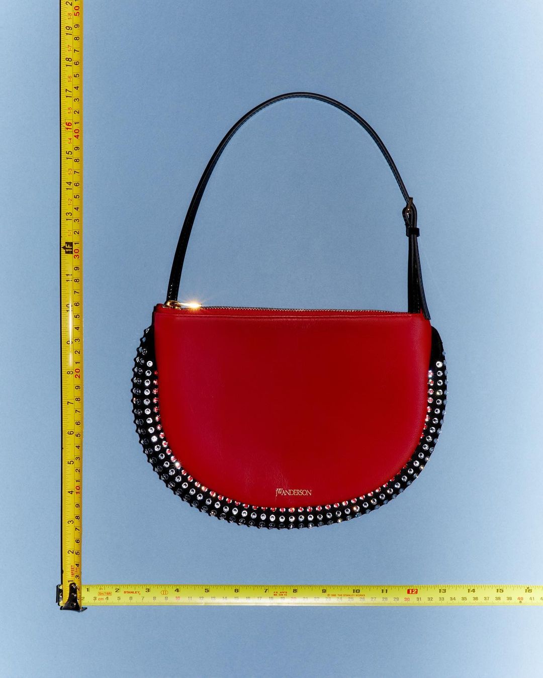Red leather JW Anderson handbag with black trim and decorative rhinestones on a blue background with yellow measuring tapes.