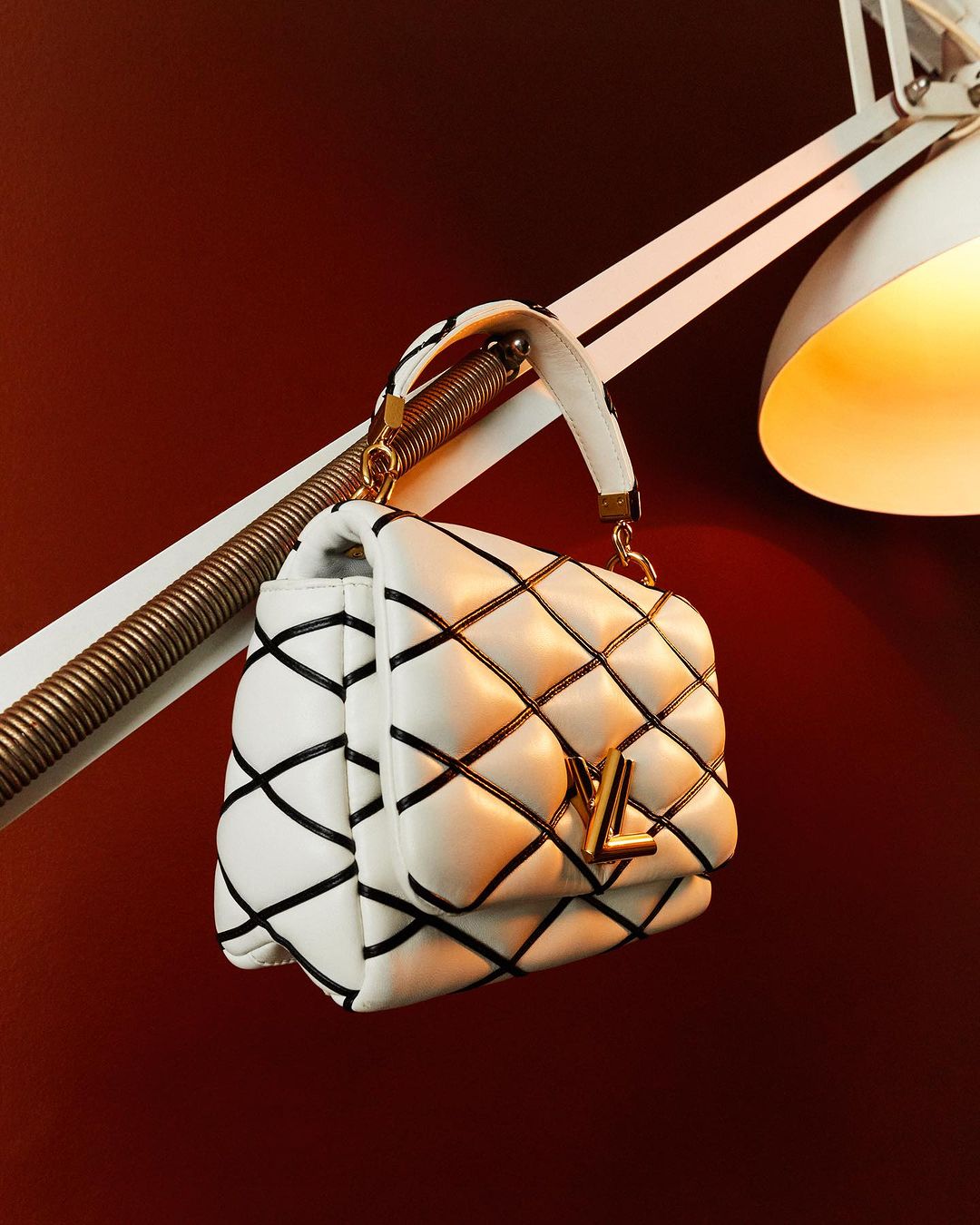 White quilted handbag with a gold logo and black trim, lit by a lamp on a dark reddish-brown background.