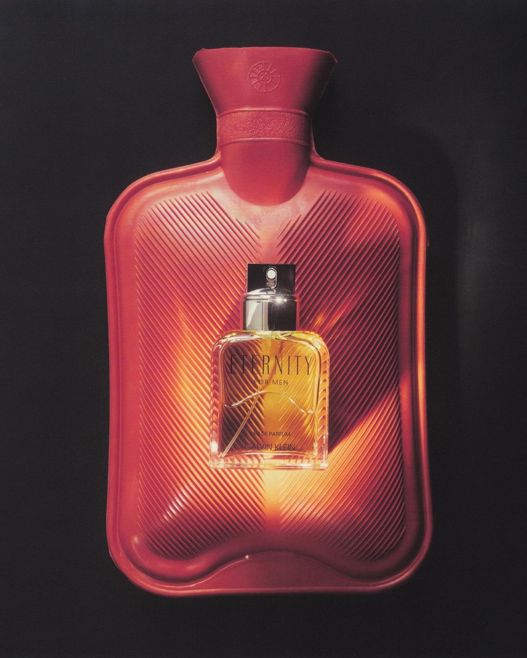 Perfume bottle centered on red hot water bottle against dark backdrop. The image highlights the scent and warmth association.