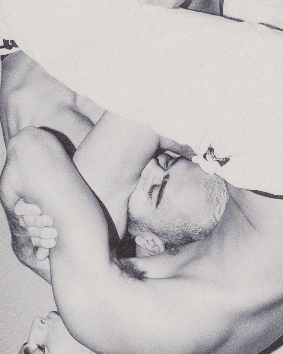Black and white image of two topless adult males in close proximity, one lying down and one in a semi-prone position with eyes closed.
