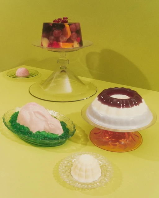 Arrangement of colorful jelly desserts on glass stands and plates against a pale green background, showcasing a variety of shapes and colors.