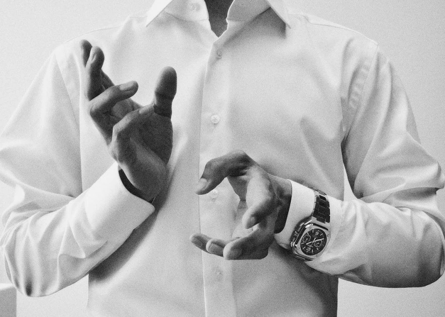 A man in a white shirt showcases a silver wristwatch with intricate detailing, captured in a monochrome, close-up shot.