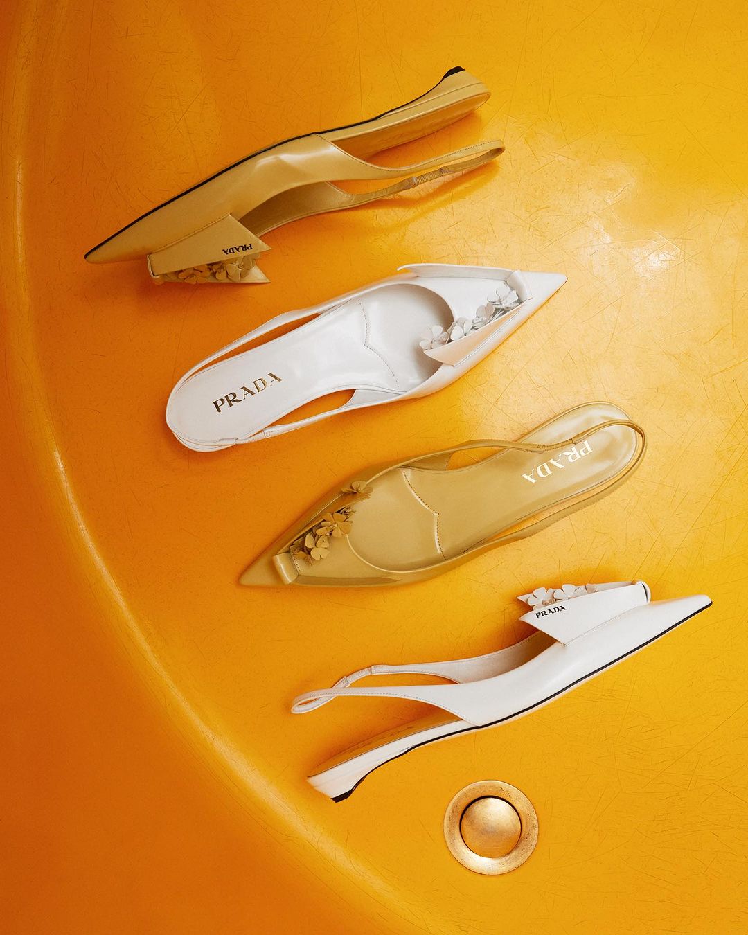 Four fashionable Prada slingback shoes in varying cream, white, and translucent colors arranged in a spiral pattern on a yellow background.