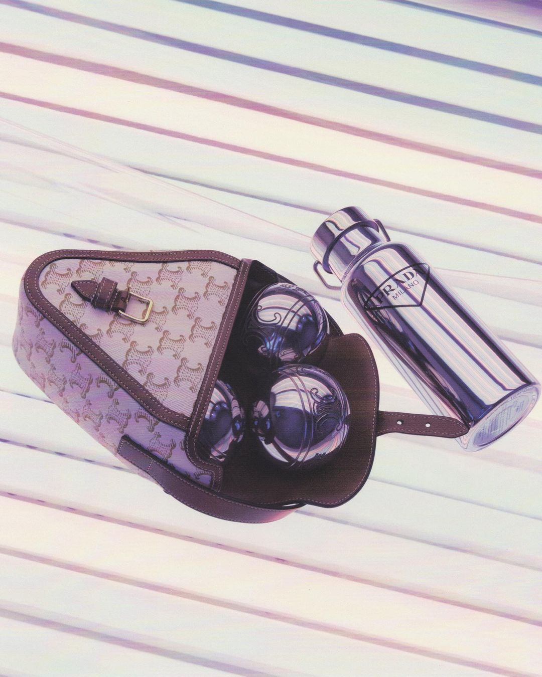 Beige purse with a brown trim filled with silver Bocce balls next to a silver Prada water bottle on a striped surface.
