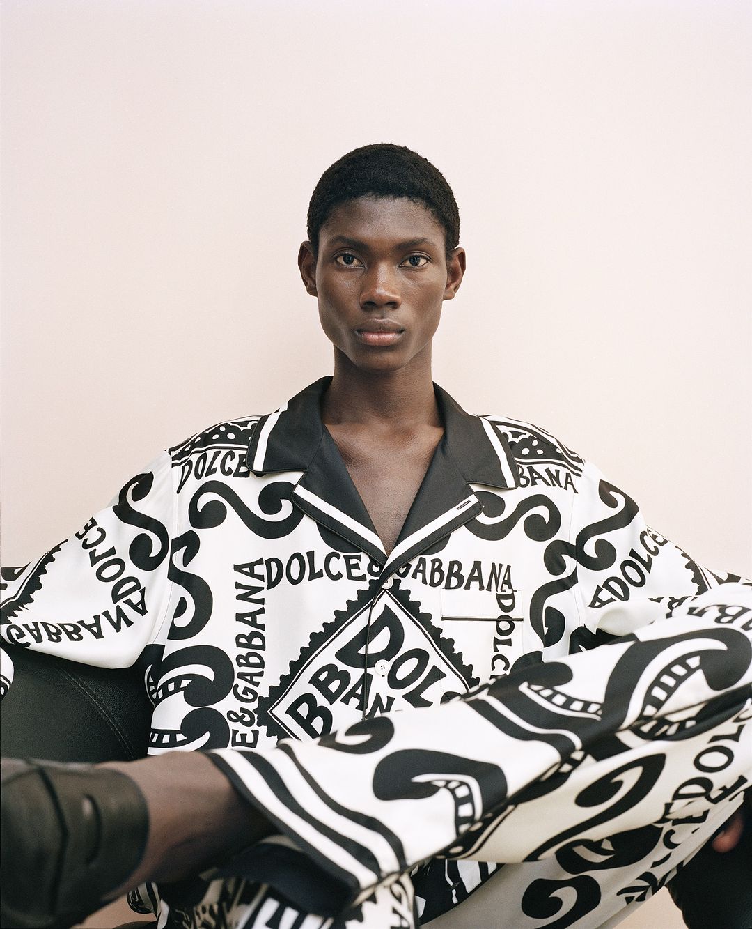 Adult Black man in a patterned Dolce & Gabbana pajama set, posing in an indoor setting with a neutral-toned background and relaxed mood.