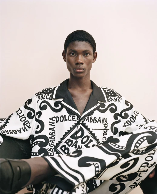 Adult Black man in a patterned Dolce & Gabbana pajama set, posing in an indoor setting with a neutral-toned background and relaxed mood.