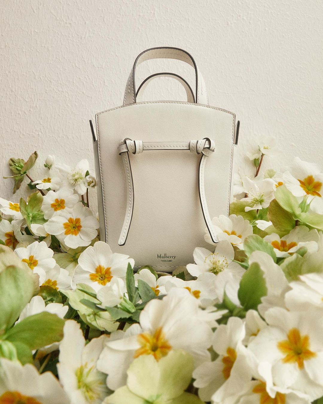 Cream-colored Mulberry purse with top handles, surrounded by primrose blossoms, against a pale backdrop. Fashion and product photography.