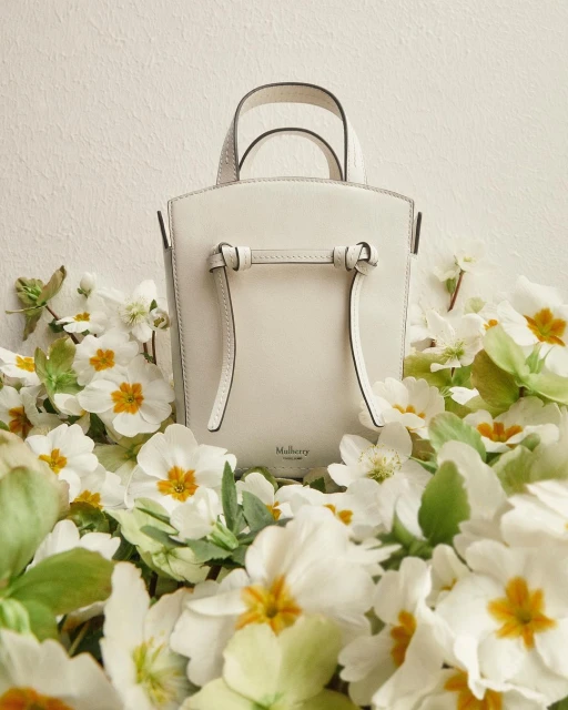 Cream-colored Mulberry purse with top handles, surrounded by primrose blossoms, against a pale backdrop. Fashion and product photography.