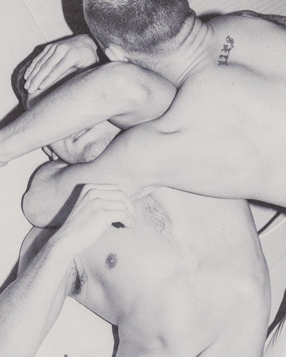 Two adult men in a close embrace, one with a tattoo on his neck, presented in a high-contrast black and white studio style photograph.