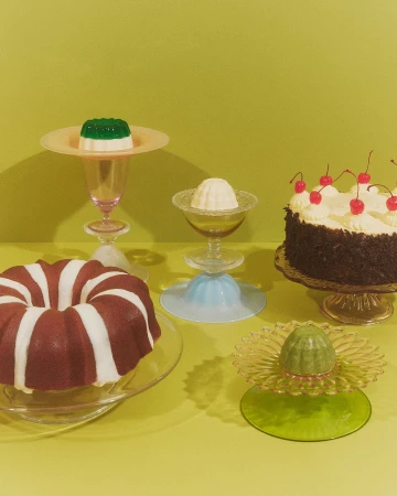 Eye-level shot of a variety of desserts on glass serving dishes against a solid light green backdrop, featuring cakes and gelatin.