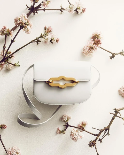 Pale blue leather purse with a gold clasp and strap, surrounded by blossoming branches on a white surface. Fashion still life.