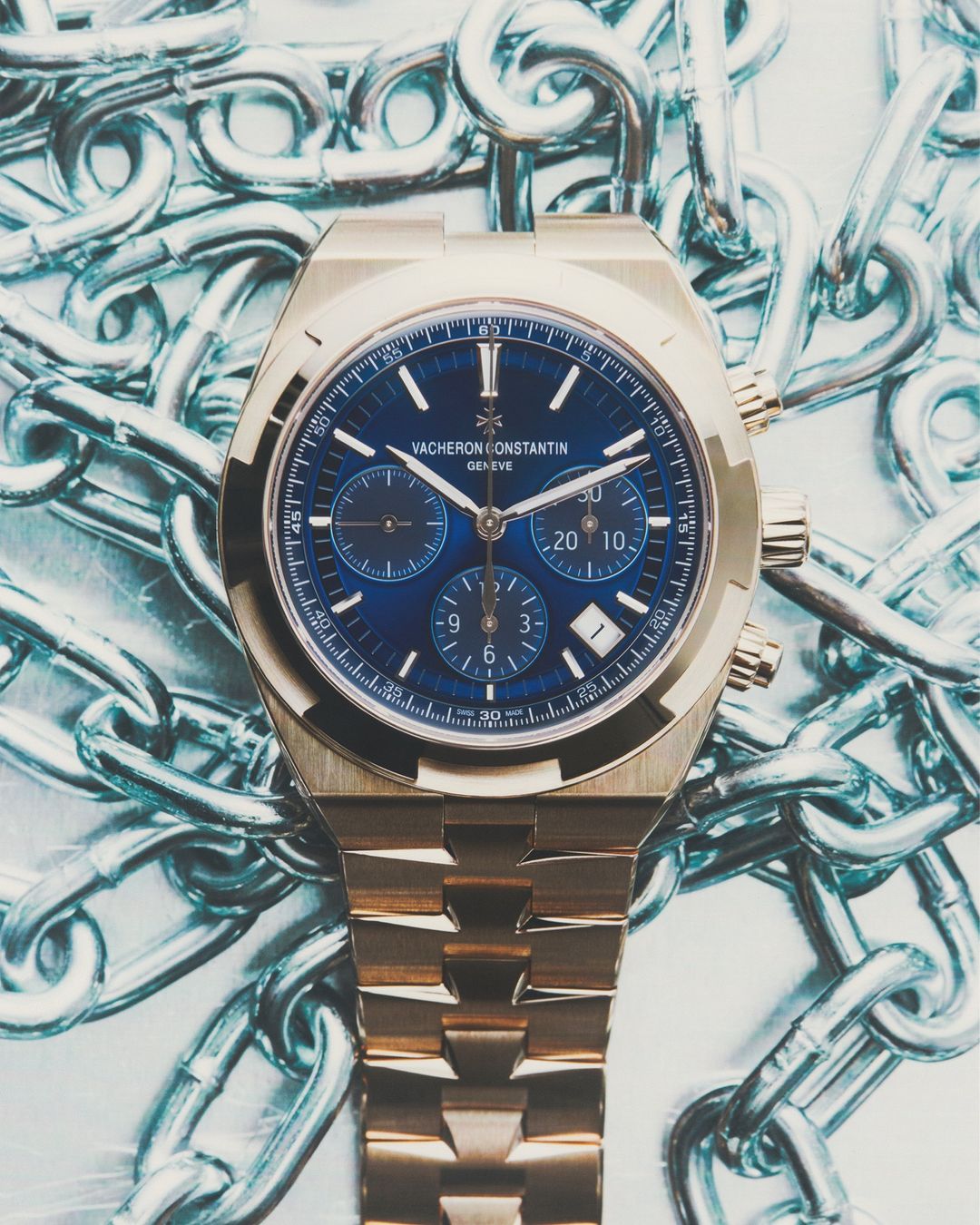 Luxury gold wristwatch with blue face is prominently displayed against a backdrop of metallic silver chains. Product photography.