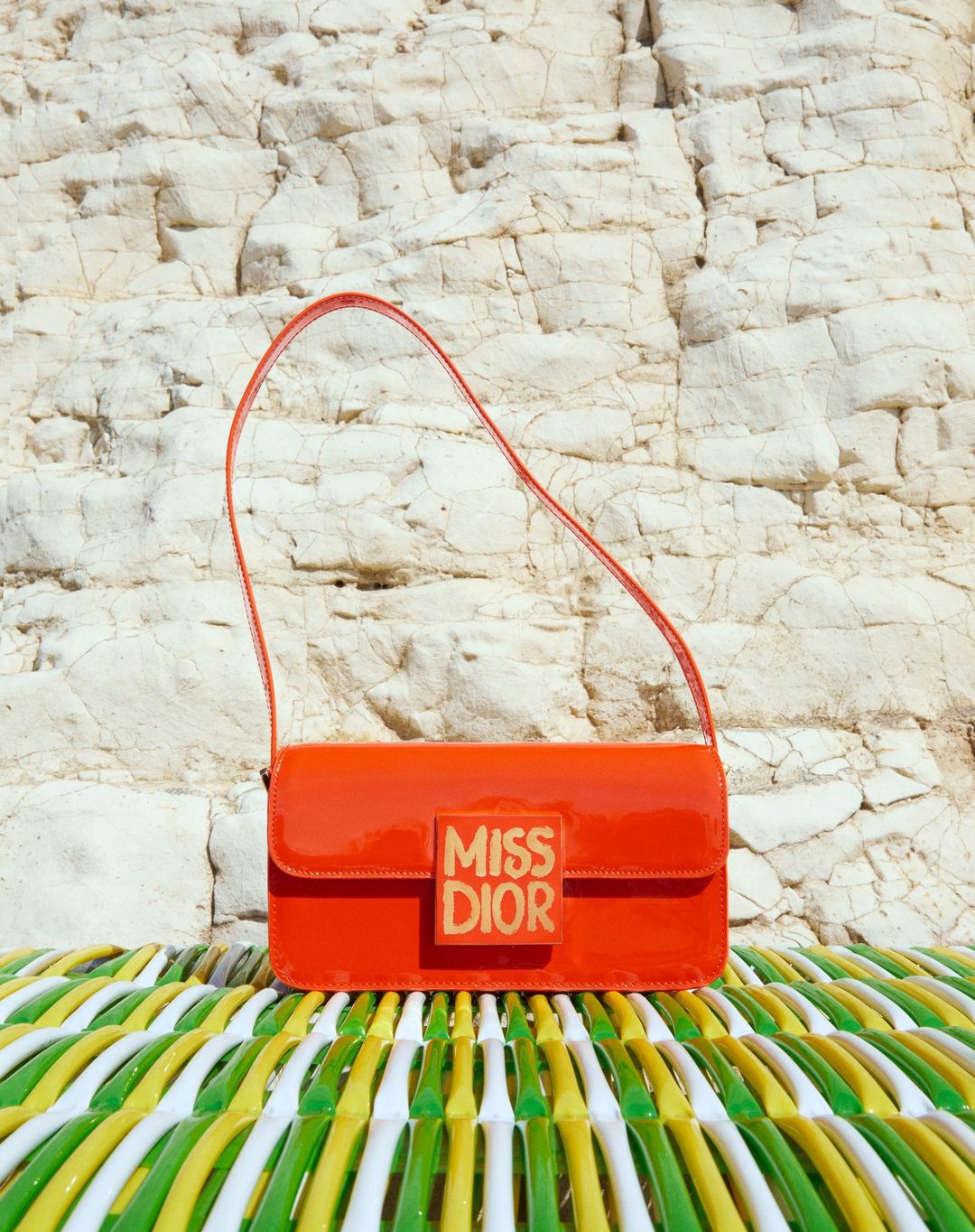 Orange Miss Dior handbag with a shoulder strap on a green and white surface in front of a textured off-white rocky background.