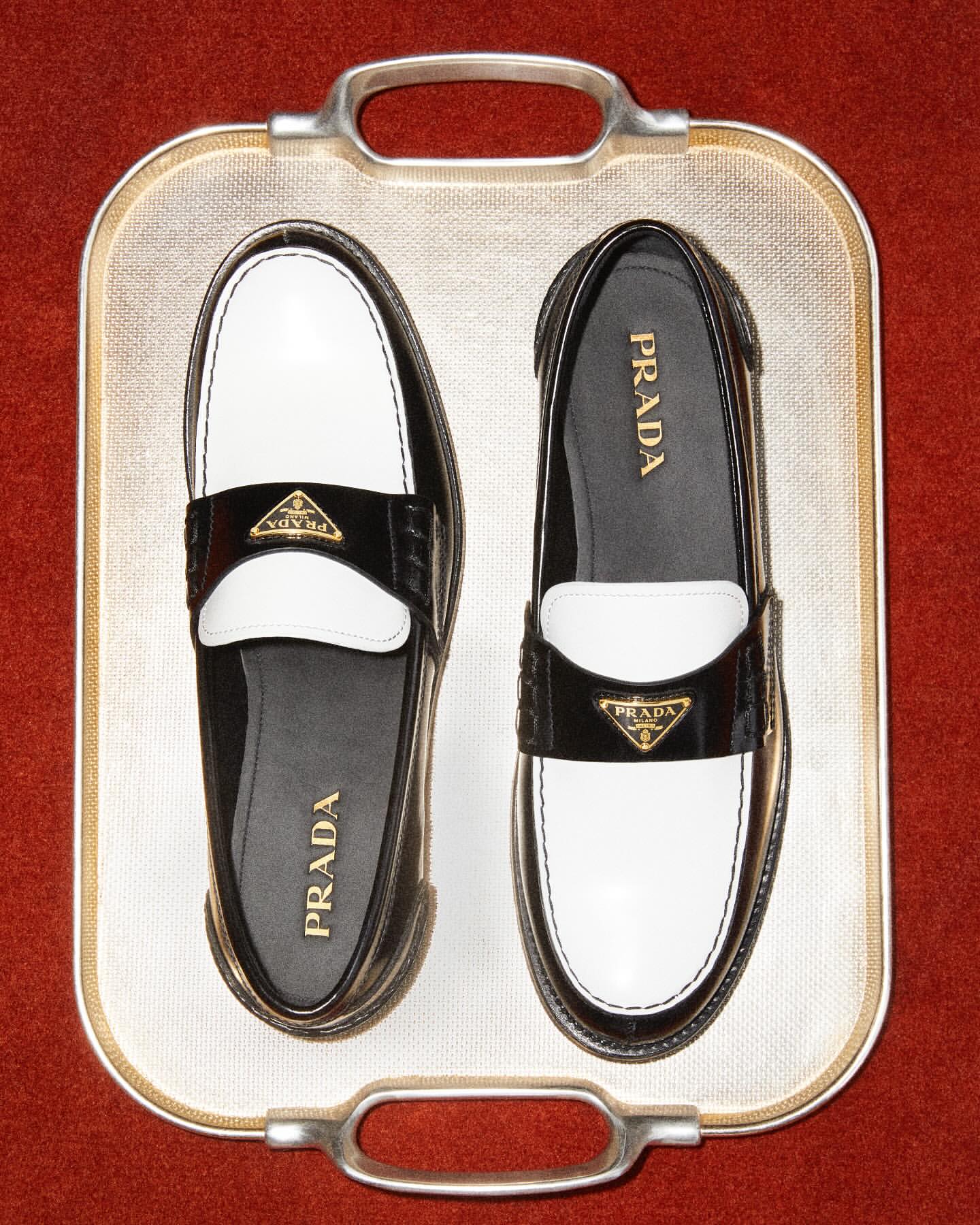 Two Prada loafers, one black/white and one white/black, are displayed on a decorative metallic tray against a red background.