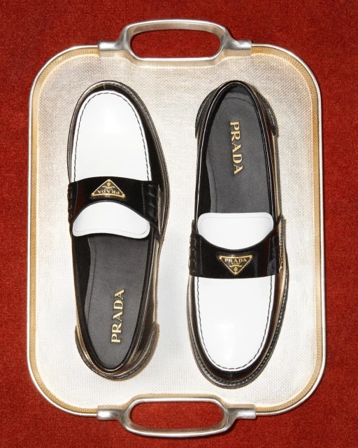 Two Prada loafers, one black/white and one white/black, are displayed on a decorative metallic tray against a red background.