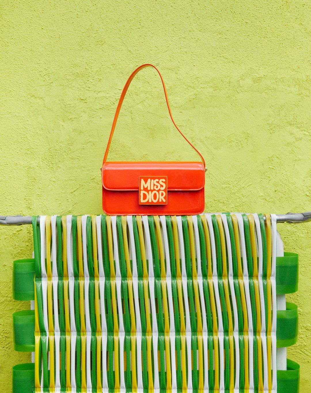 Bright orange Miss Dior handbag with logo displayed on a green surface with white, green, and yellow striped design feature.