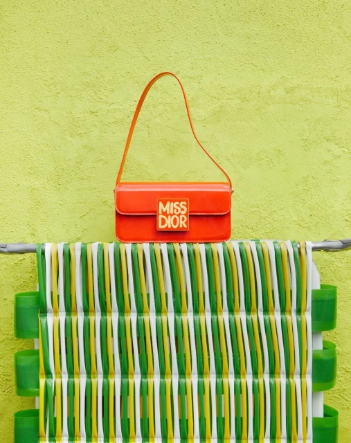 Bright orange Miss Dior handbag with logo displayed on a green surface with white, green, and yellow striped design feature.
